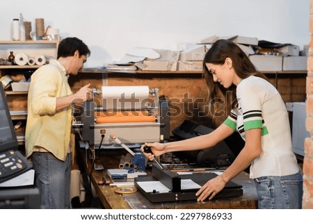 cheerful typographer using paper trimmer near colleague next to professional print plotter