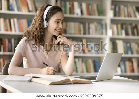 Cheerful university student girl working on research study in library, using headphones, laptop for communication, video call, watching learning webinar, online seminar, writing