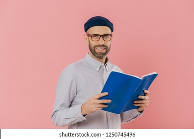 Cheerful unshaven clever man holds book in front reads exciting story, studies scientific literature, wears glasses and white shirt, stands against pink background. People, reading, spare time concept - Shutterstock ID 1250745496