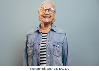 Cheerful Very Emotional Old Woman With Gray Hair Wearing Jeans Jacket And Shirt Looking At Camera, Laughing And Posing Isolated Over Blue Background. Smart Happy Senior Lady. Happy Feelings Concept