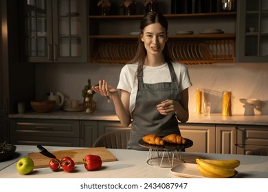 A cheerful video blogger stands in a well-equiped, modern kitchen presenting freshly baked croissants on a cooling rack. She wears a gray apron over casual clothing, and the kitchen counter is neatly