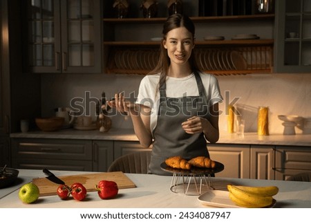 A cheerful video blogger stands in a well-equiped, modern kitchen presenting freshly baked croissants on a cooling rack. She wears a gray apron over casual clothing, and the kitchen counter is neatly