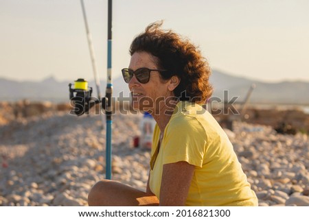 A cheerful and vital woman, in her 70s, fishing with her fishing rod, on a natural beach, at sunset, next to the Mediterranean Sea, Spain