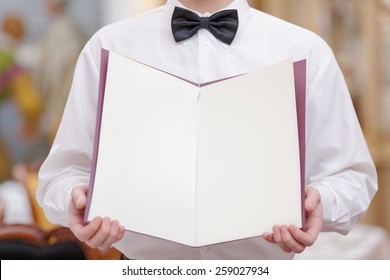 Cheerful Waiter. Cropped Image Of Handsome Young Waiter In Shirt And Bow Tie Holding A Menu With Blank Pages For Copy Space In The Luxury Restaurant