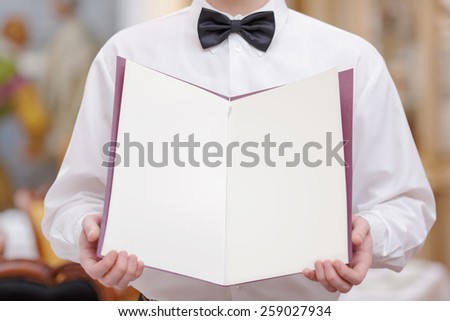 Cheerful waiter. Cropped image of handsome young waiter in shirt and bow tie holding a menu with blank pages for copy space in the luxury restaurant