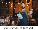 A cheerful waiter welcomes guests while holding a tablet in a warm, stylish restaurant. The atmosphere is inviting and modern, with people enjoying their meals and conversations.