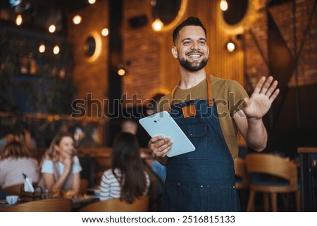 A cheerful waiter welcomes guests while holding a tablet in a warm, stylish restaurant. The atmosphere is inviting and modern, with people enjoying their meals and conversations.