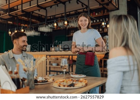 Cheerful waitress laughing while taking orders from customers sitting at a table with pizza in a pizzeria restaurant