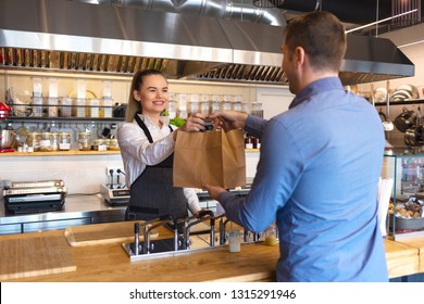 Cheerful waitress wearing apron serving customer at counter in restaurant - Small business and service concept with young business owner woman giving bag with takeaway food to client - Shutterstock ID 1315291946