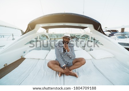 A cheerful wealthy bearded mature black guy in an elegant striped black and white costume with shorts and hat is sitting outdoors on the deck of a luxury bright speed yacht moored on a berth