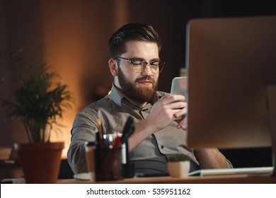 Cheerful Web Designer Dressed In Shirt And Wearing Eyeglasses Working Late At Night And Chatting By Phone.