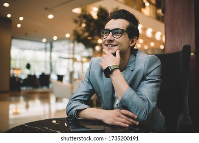 Cheerful Well Dressed Male In Eyeglasses Looking Up And Smiling While Sitting At Table With Mobile Phone In Luxury Restaurant