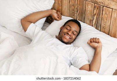 Cheerful Wellslept African Man Awakening Stretching Hands Waking Up Lying In Modern Bedroom At Home, Smiling With Eyes Closed. Rested Guy Enjoying Good Morning In Bed. Above View