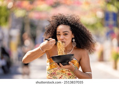 A cheerful woman of African or Afro-Latin ethnicity, wearing a vibrant outfit, enjoys eating noodles outdoors. She smiles brightly while holding a bowl, capturing a fun and relaxed moment.