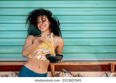 A cheerful woman of African or Afro-Latin ethnicity, wearing a vibrant outfit, enjoys eating noodles outdoors. She smiles brightly while holding a bowl, capturing a fun and relaxed moment.