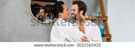 cheerful woman applying face cream on nose of smiling boyfriend in bathrobe, banner