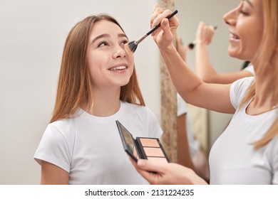 Cheerful woman applying powder on face of smiling teenage girl while doing makeup at home