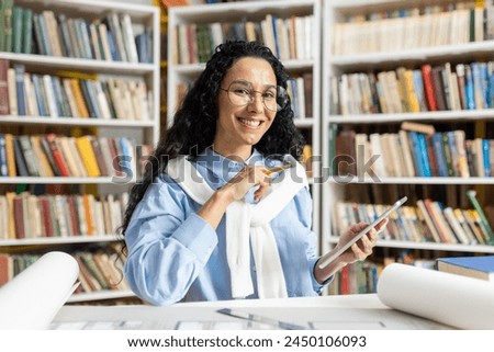 A cheerful woman in a blue shirt and white scarf, using her smartphone and smiling, seated in a well-stocked library. She exudes an enthusiastic and studious vibe.