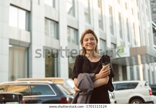 Cheerful woman with\
broad smile holding coffee and jacket walking near the office\
building and car parking. Smiling young lady feeling happy\
strolling down the urban\
street.