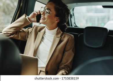 Cheerful woman in business suit sitting on backseat of her car and talking on mobile phone. Businesswoman using phone while traveling by a car.