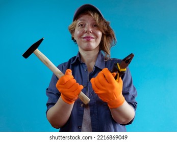 A Cheerful Woman In A Cap And Shirt Holds A Hammer And Pliers In Her Hand. Portrait Of A Woman In Orange Gloves. Foreman Woman. Woman Plumber