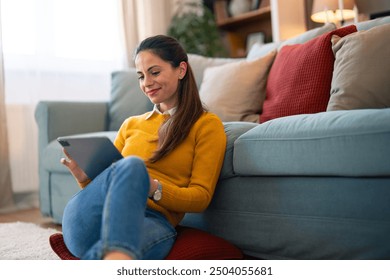 A cheerful woman in casual attire enjoys browsing on her digital tablet while comfortably seated on the floor against a couch in a well-lit living room. - Powered by Shutterstock