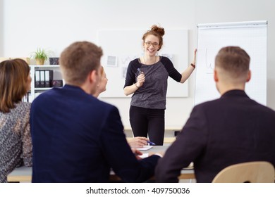 Cheerful Woman In Casual Wear And Eyeglasses At Large Flip Chart During Staff Meeting With Male And Female Co-workers