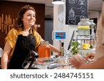 Cheerful woman at checkout counter in zero waste shop selling healthy lentils in jar to customer with green living lifestyle. Client buys food in local neighborhood store from smiling vendor