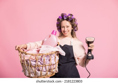 A Cheerful Woman Cleaning People's Homes Working As A Cleaner. Domestic Servant Inserts Laundry, Irons Clothes In Apron With Rollers On Head Portrait On Pink Background.