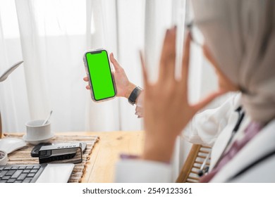 A cheerful woman in a colorful hijab holds a smartphone with a green screen at home - Powered by Shutterstock