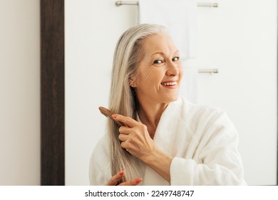 Cheerful woman combing her long hair with a wood comb in front of a bathroom mirror - Powered by Shutterstock