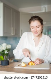 Cheerful Woman Is Cooking In Her Smart Kitchen With A Portable Speaker And Her Voice Recipe. Cute Chef Cuts Apples To The Sounds Of The Personal Audio Assistant