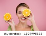 cheerful woman covering eye with half of ripe orange isolated on pink