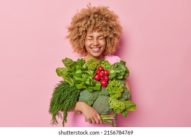 Cheerful woman with curly hair carries different fresh vegetables keeps eyes closed smiles broadly isolated over pink background. Healthy organic food and spring harvest concept. Garden produce