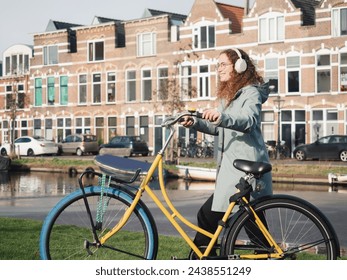 A cheerful woman with curly hair and headphones enjoys a sunny day, walking with her yellow bicycle by a canal in Amsterdam, with classic Dutch houses in the background - Powered by Shutterstock