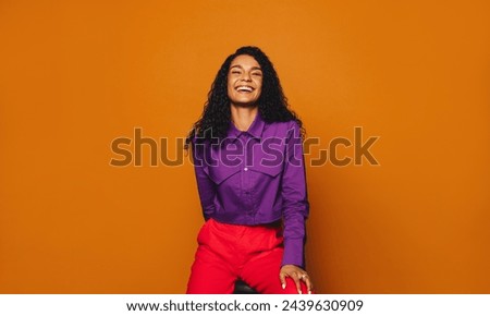 Cheerful woman with curly hair sits in casual clothing, smiling at the camera. Her vibrant, stylish fashion sense stands out against the orange background.
