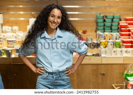 A cheerful woman with curly hair stands hands on hips in a cozy pottery and home decor store, exuding confidence and positivity.