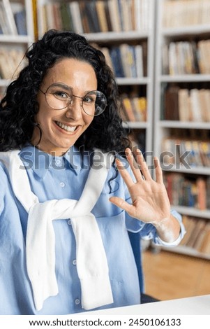 A cheerful woman with curly hair, wearing glasses and a casual outfit, happily waves in a well-stocked library, surrounded by books.