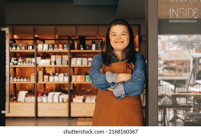 Cheerful Woman With Down Syndrome Standing At The Entrance Of A Deli. Empowered Woman With An Intellectual Disability Working As A Shopkeeper In A Local Supermarket.