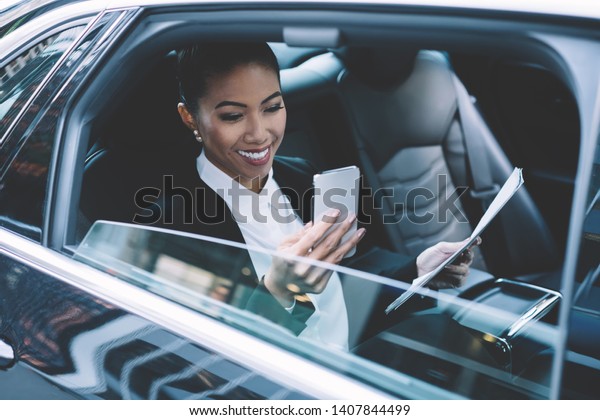 Cheerful woman in formal wear holding contract\
documents in hand and smiling from received email on modern cell\
smartphone device, happy female manager spending time for chatting\
on way to office
