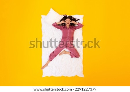 Cheerful Woman Gesturing Peace Signs Lying On Blanket Wearing Pajamas Smiling To Camera Over Yellow Studio Background, Top View Shot. Female Having Fun Posing And Relaxing At Bedtime