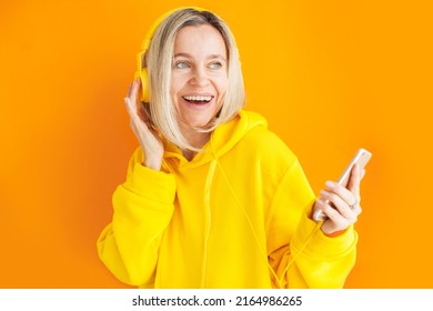 Cheerful Woman Girl In Yellow Hoody Posing Isolated On Yellow Wall Background Studio Portrait. Middle Aged People Lifestyle Concept. Mock Up Copy Space. Listen Music With Headphones And Phone, Dancing