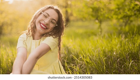 A cheerful woman with glasses sitting in a grassy field, smiling brightly under the sunshine. Captures happiness, relaxation, and natural beauty. - Powered by Shutterstock