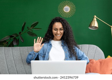 Cheerful woman in glasses waving during a video call on laptop at home, expressing joy and friendliness in a casual setting. - Powered by Shutterstock