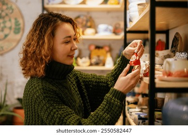 A cheerful woman in a green knit sweater selects a handmade ceramic Christmas decoration in a festive artisan shop. The image highlights the charm of local craftsmanship and holiday spirit - Powered by Shutterstock