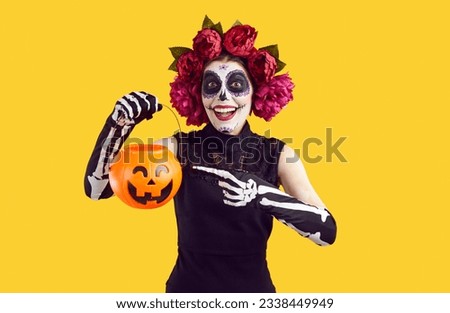 Cheerful woman in Halloween Mexican costume hints with gesture that she wants to trick or treat. Woman on orange background points with her finger to the pumpkin candy basket in her hand.