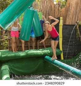 Cheerful woman having fun with friends in summer amusement park, trying to walk on inflatable log above pool and not to fall into water between swinging obstacles knocking player down - Powered by Shutterstock