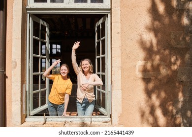 Cheerful woman and her daughter waving to somebody while looking from the window - Shutterstock ID 2165441439