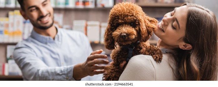 Cheerful woman holding brown poodle near blurred muslim salesman in pet shop, banner  - Shutterstock ID 2194663541