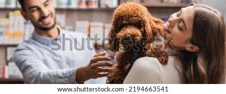 Cheerful woman holding brown poodle near blurred muslim salesman in pet shop, banner 